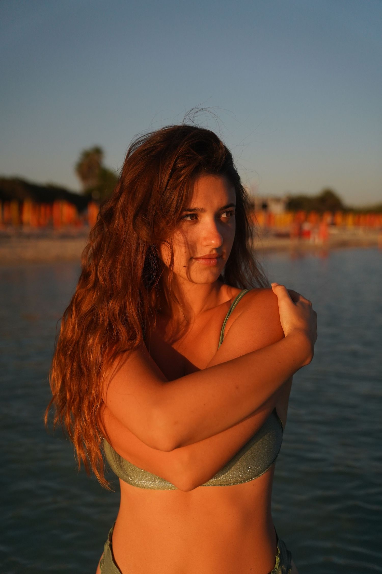 Woman in sage swimwear poses at beach during sunset with arms crossed.
