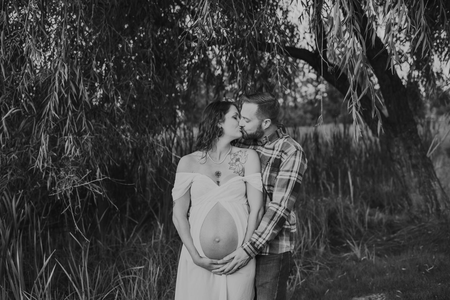 Black and white maternity photo under willow tree branches in outdoor setting.