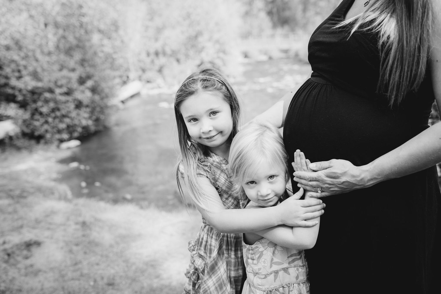 Black and white photo of children hugging pregnant belly outdoors.