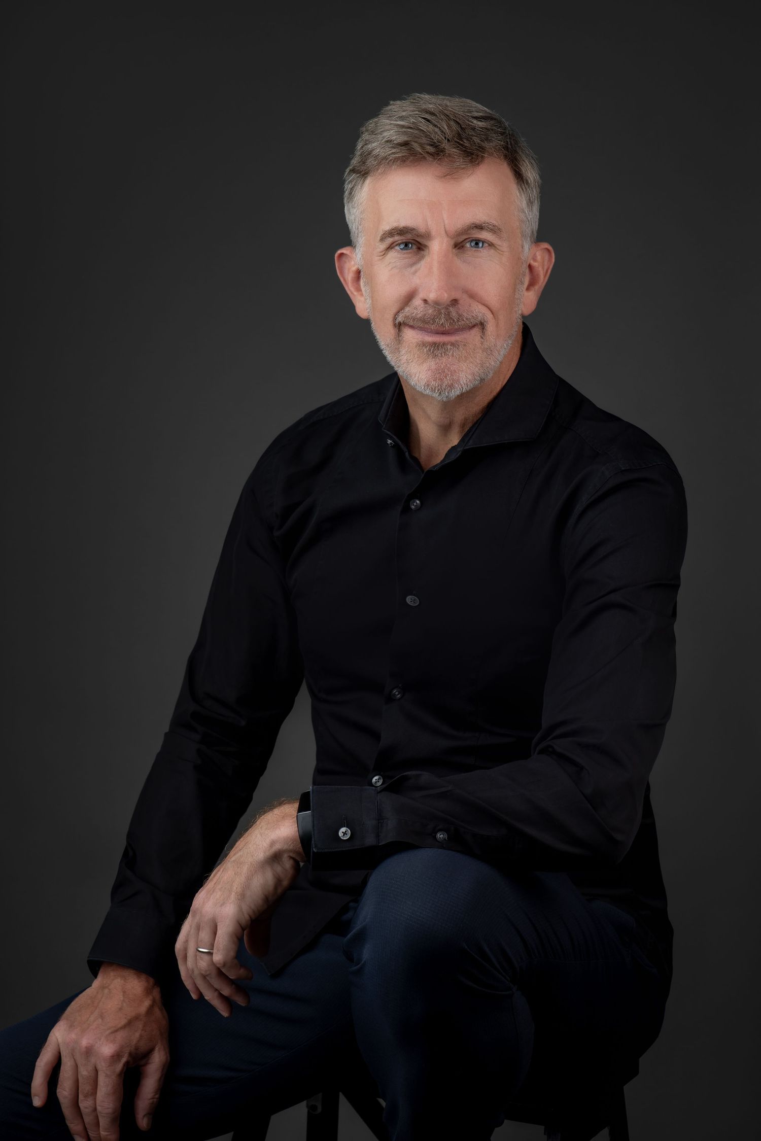 A professional portrait of a man with short gray hair, dressed in a black shirt, seated confidently against a dark studio background.