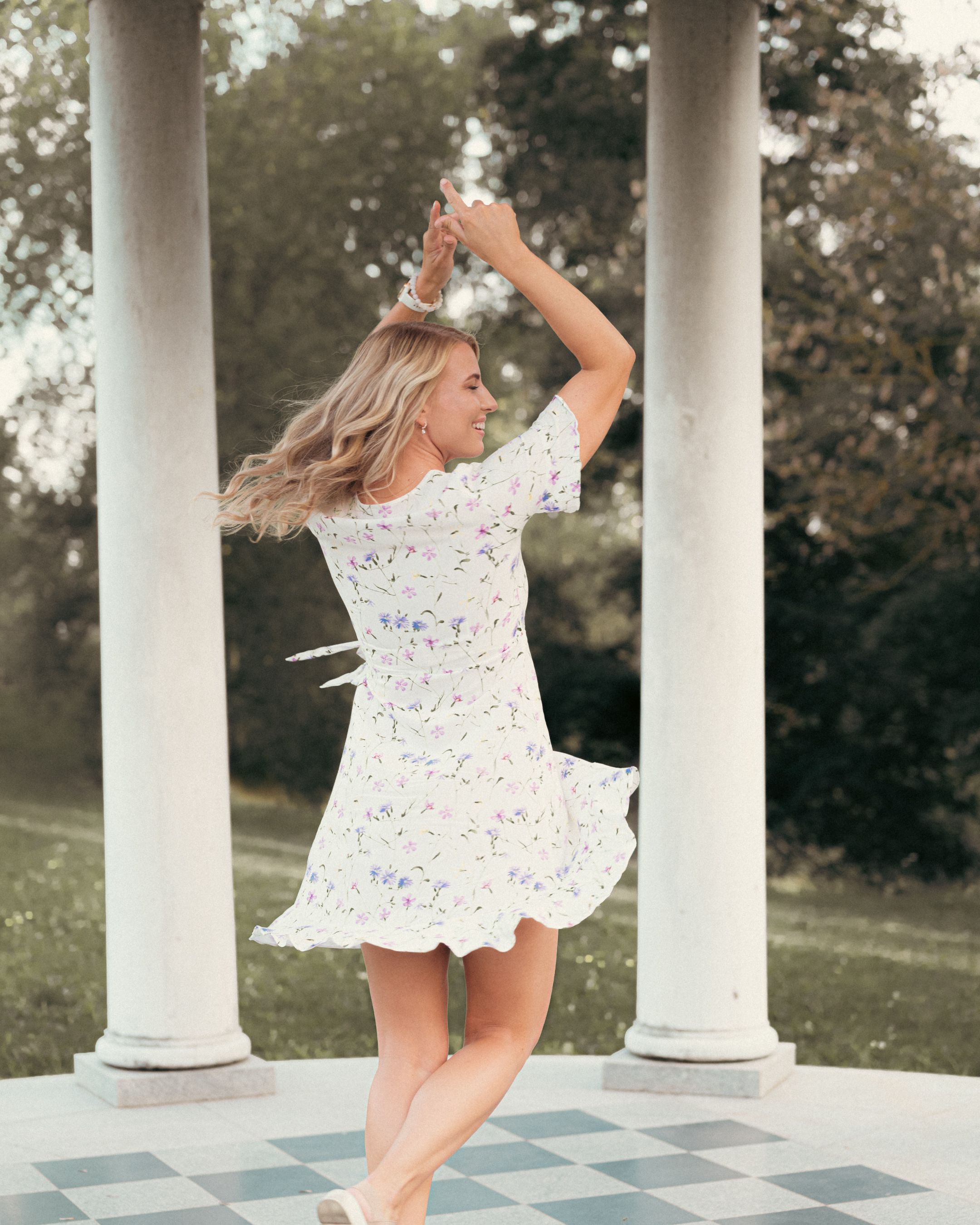 Woman in white floral dress dances between classical white columns on checkerboard floor.