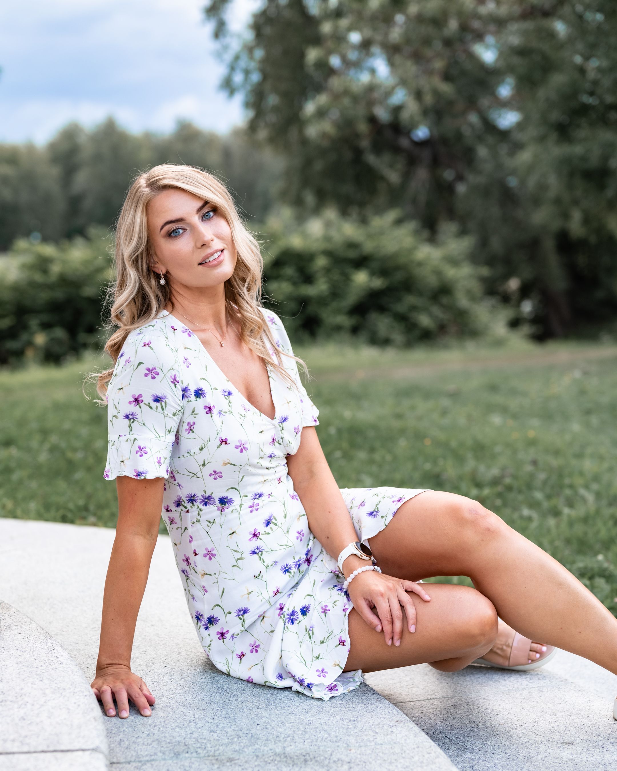 Woman in floral summer dress poses on concrete ledge in outdoor garden setting.