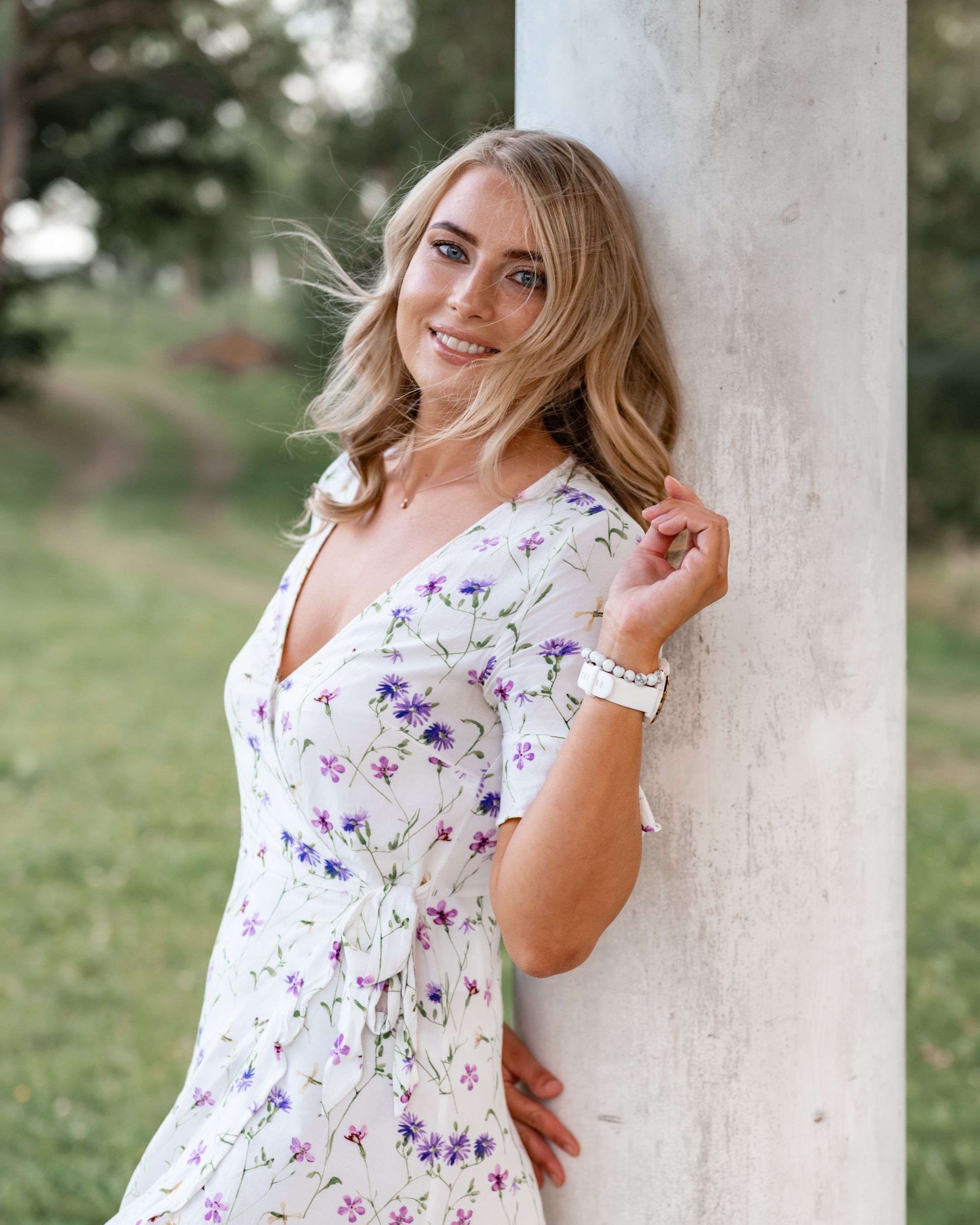 Woman in purple floral dress poses against white column in natural garden setting.