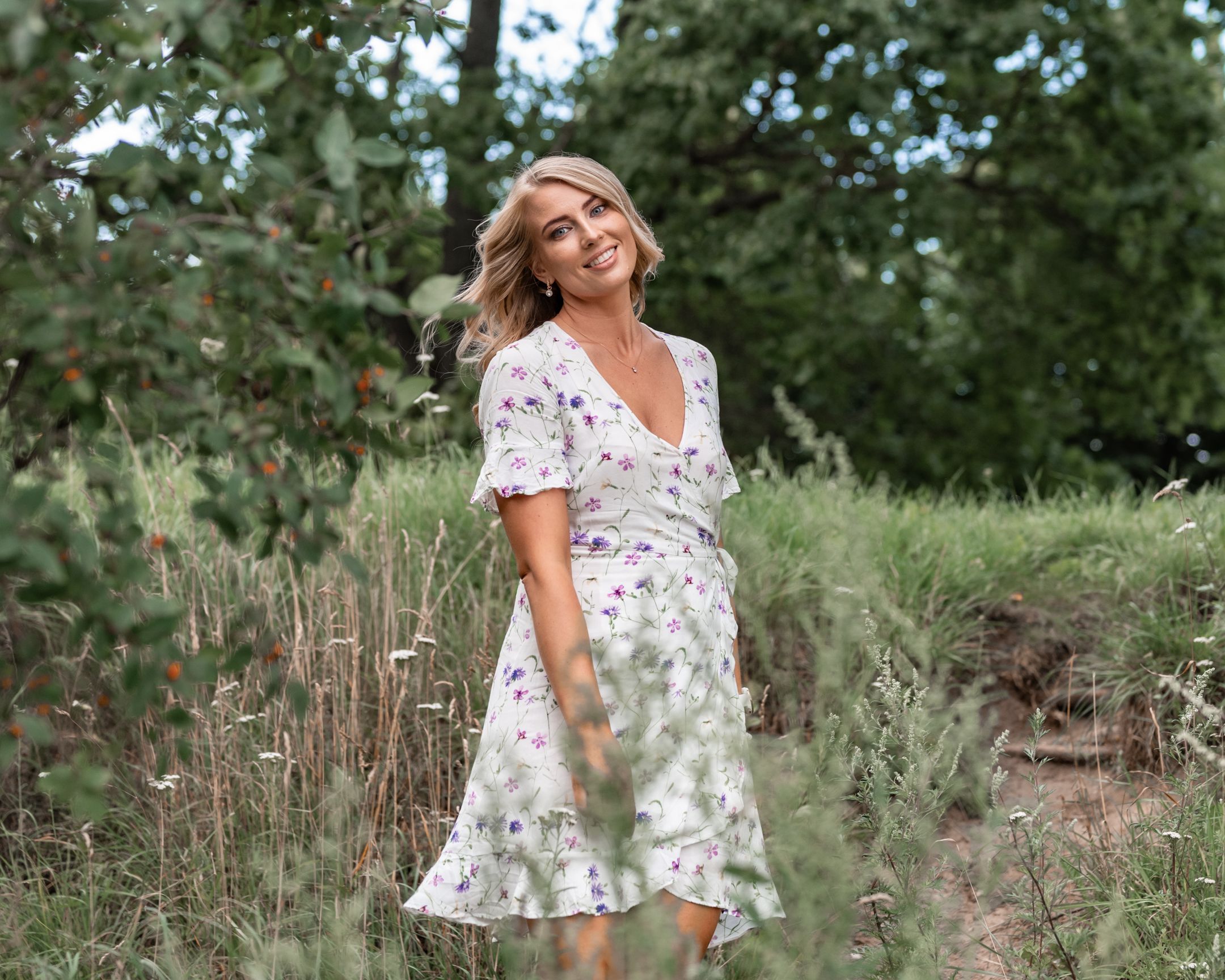 Woman in floral dress stands among tall grasses in natural outdoor setting.