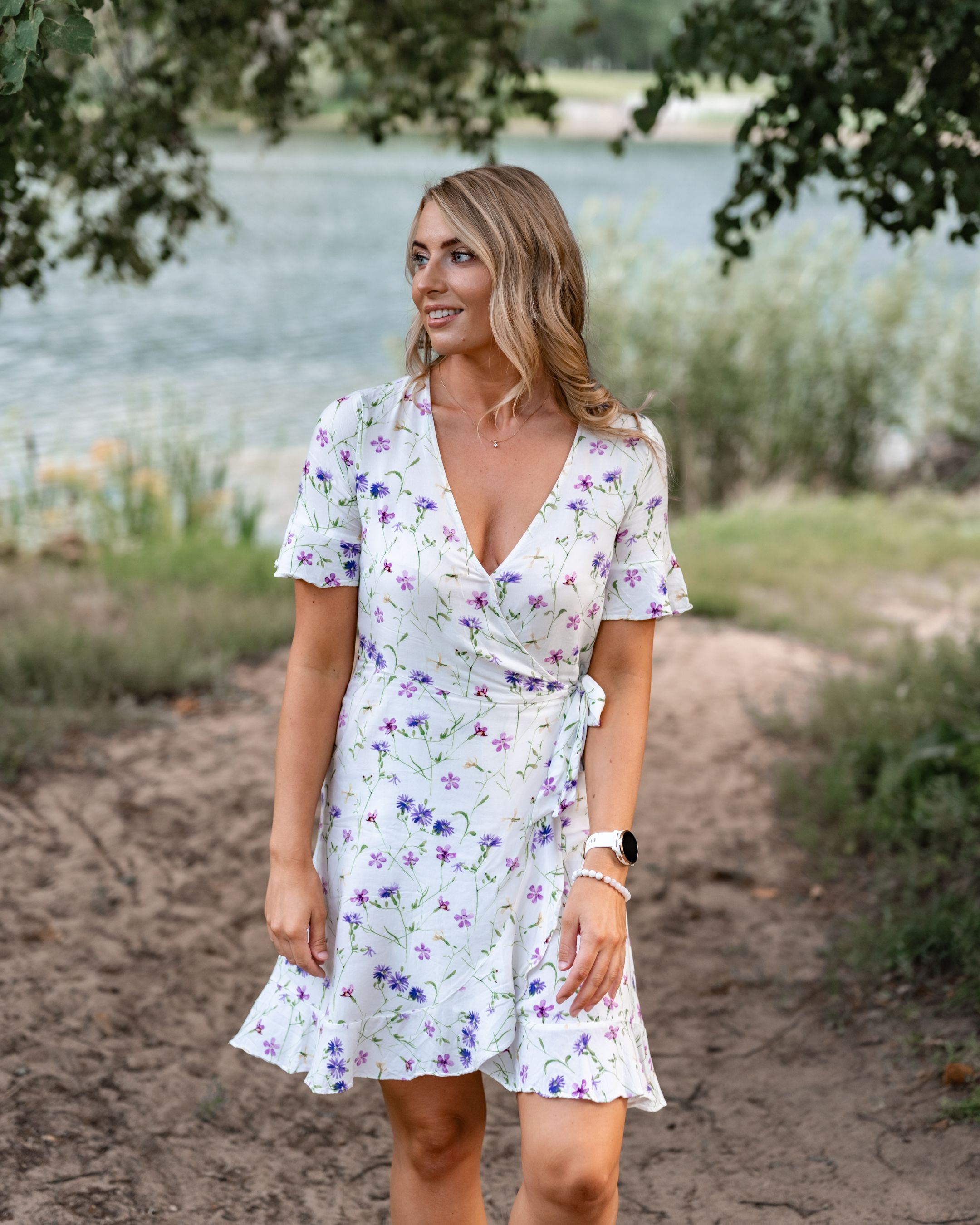 A white floral wrap dress with purple details is modeled outdoors near a lake.