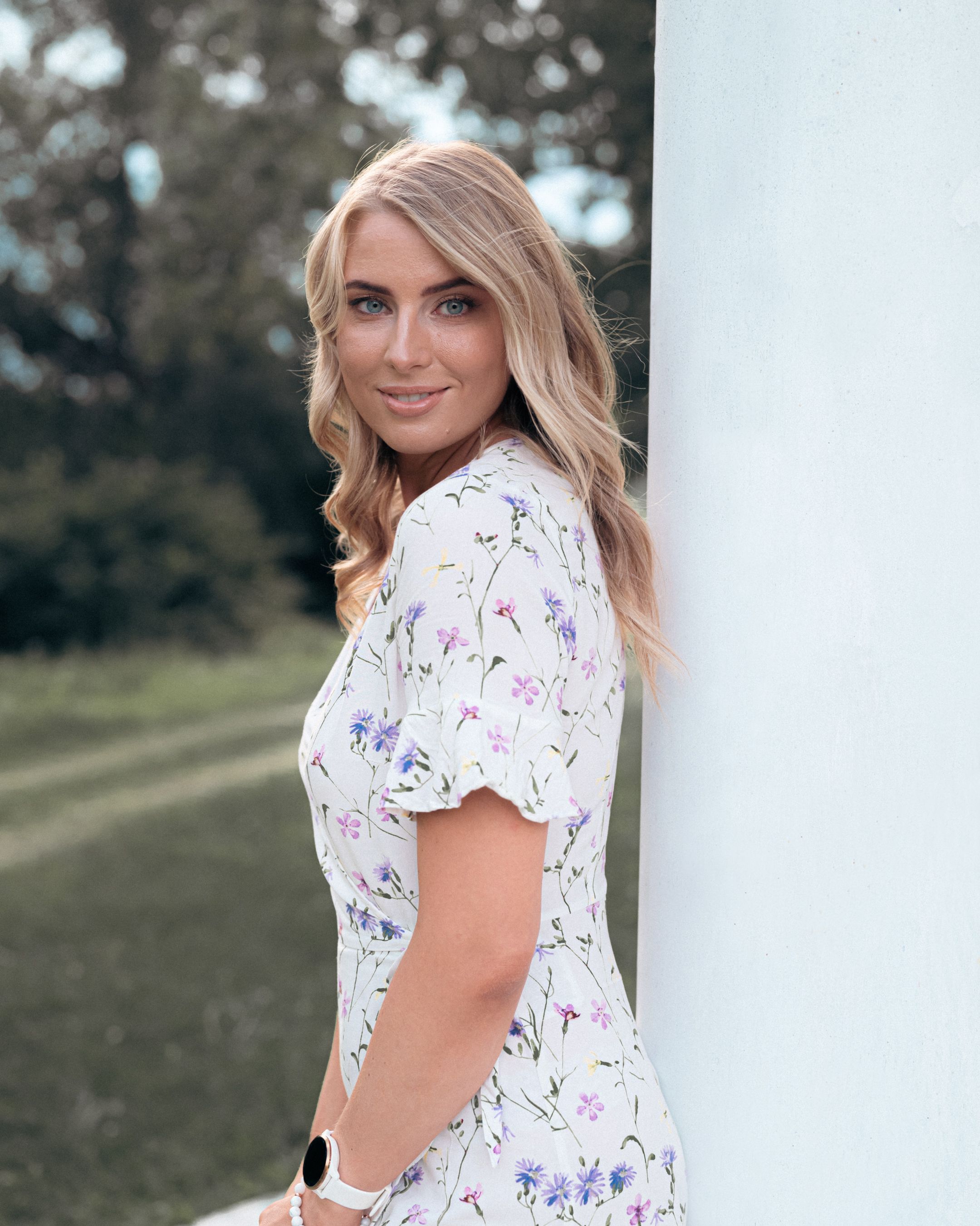 Woman in a white floral dress poses outdoors with soft lighting and natural scenery in the background.