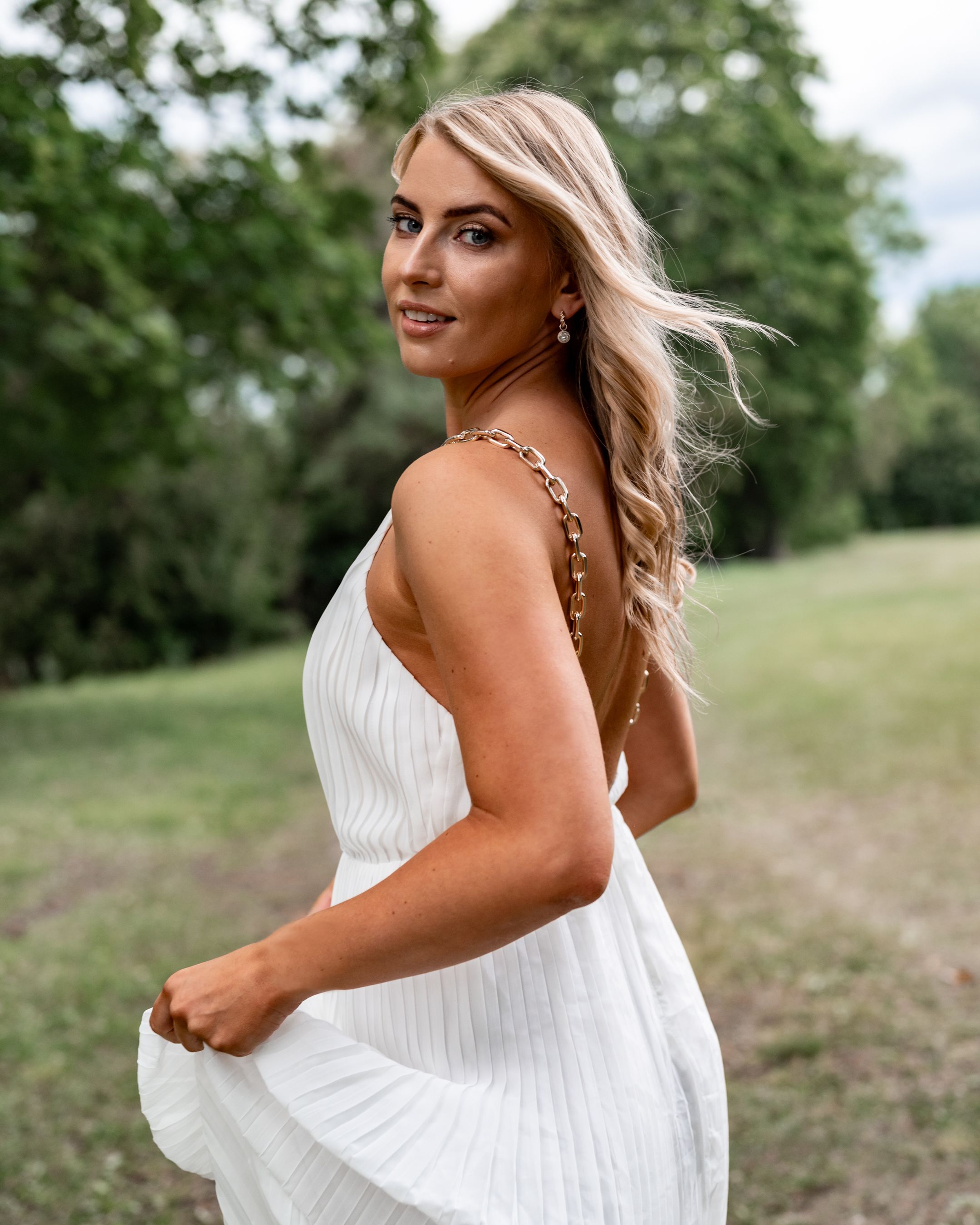 Woman in white pleated dress twirls gracefully in an outdoor garden setting.