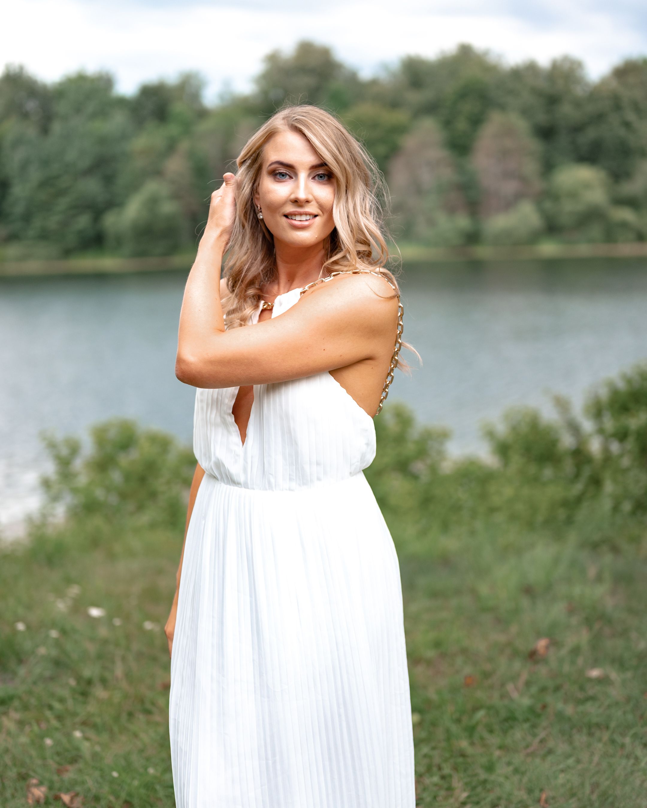 Portrait of woman in white sundress standing by a scenic lake with trees in the background.