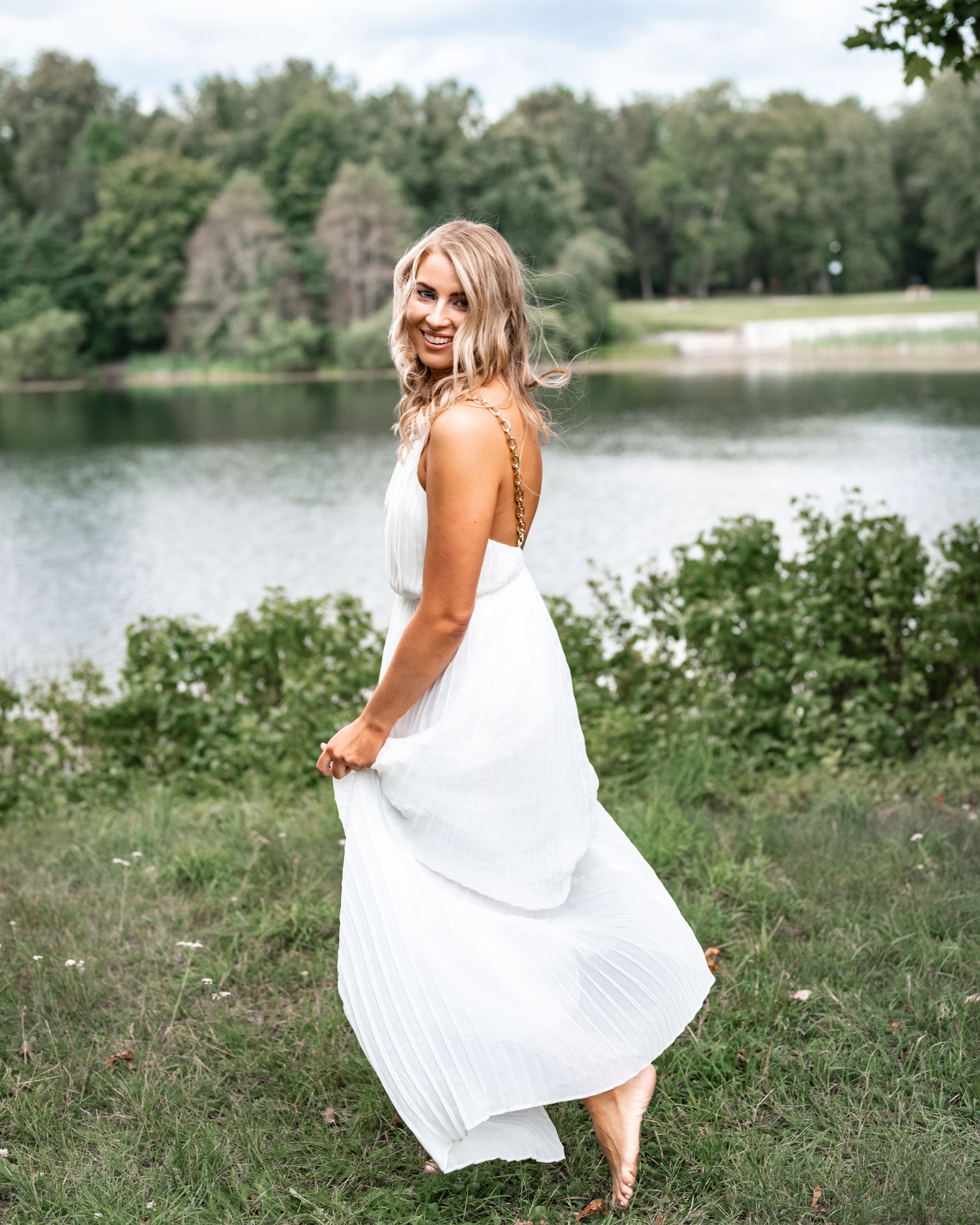 Woman in flowing white dress twirls by a serene lake surrounded by trees.