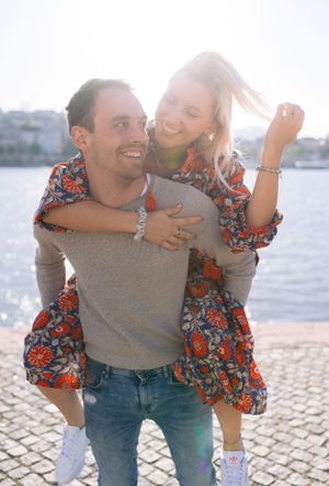 A joyful couple poses by the waterfront, the man giving the woman a piggyback ride in matching floral outfits.