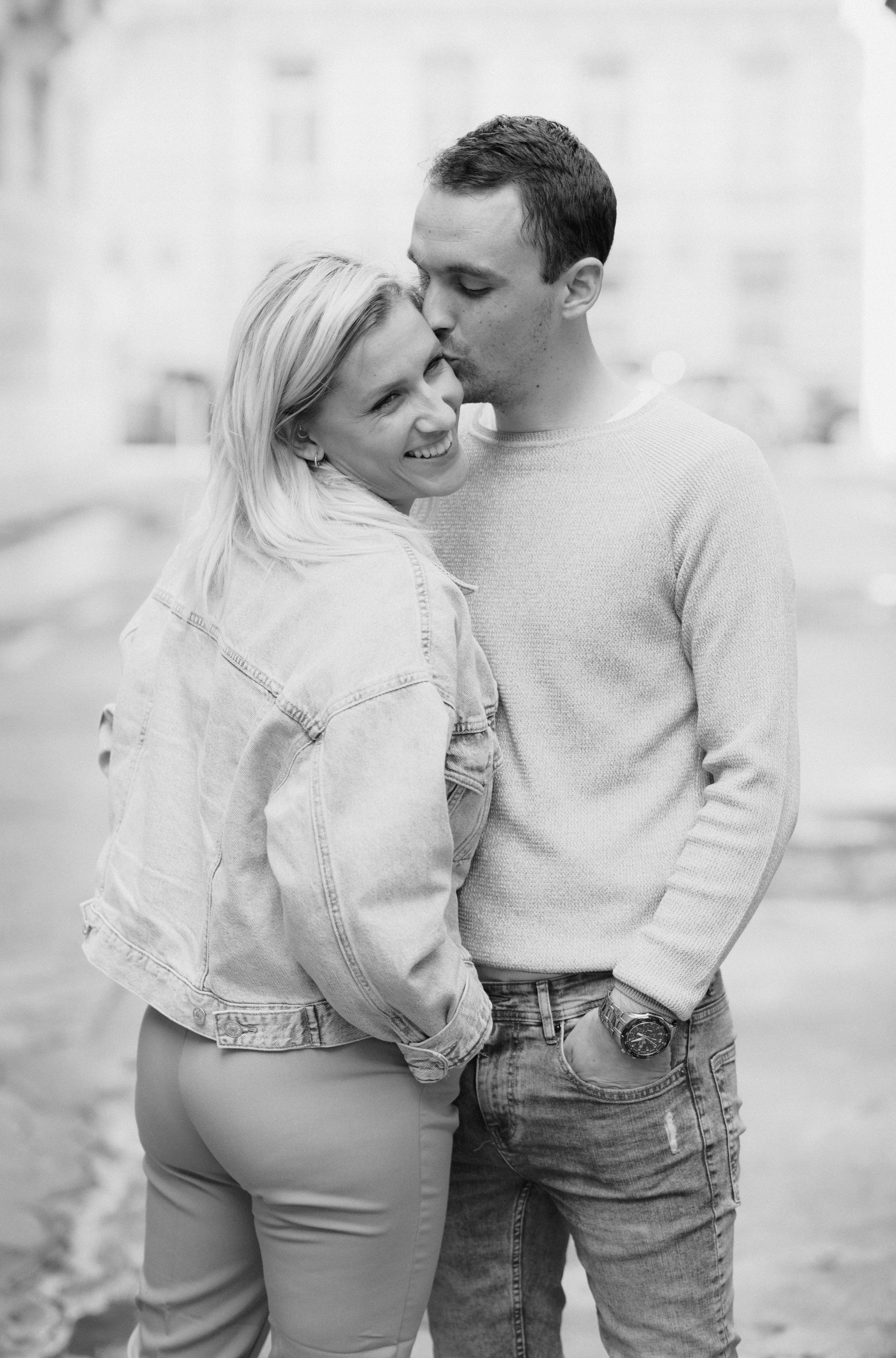A black and white photo of a couple sharing a kiss outdoors, the man leaning in while the woman smiles.