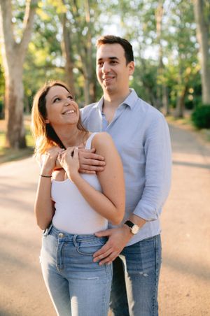 Smiling couple embracing on a tree-lined path in a sunny park, woman looking up joyfully.