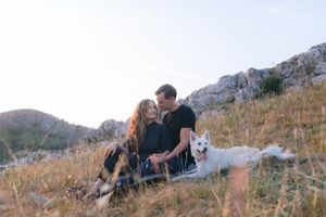 Romantic couple sitting in a grassy field kissing with a white dog beside them and rocky hills in the background.