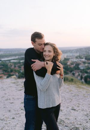 A couple embraces on a hilltop at sunset, the man hugging the woman from behind as she smiles warmly.
