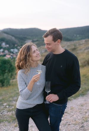 Happy couple holding champagne glasses smiling at each other outdoors with scenic hills at sunset.