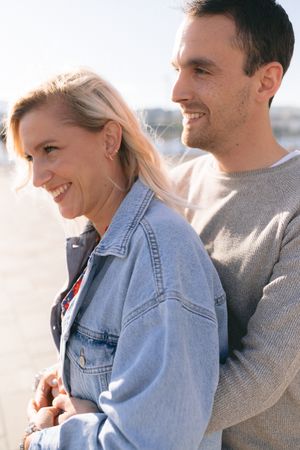 A smiling couple stands close together outdoors in bright sunlight, the woman in a denim jacket.