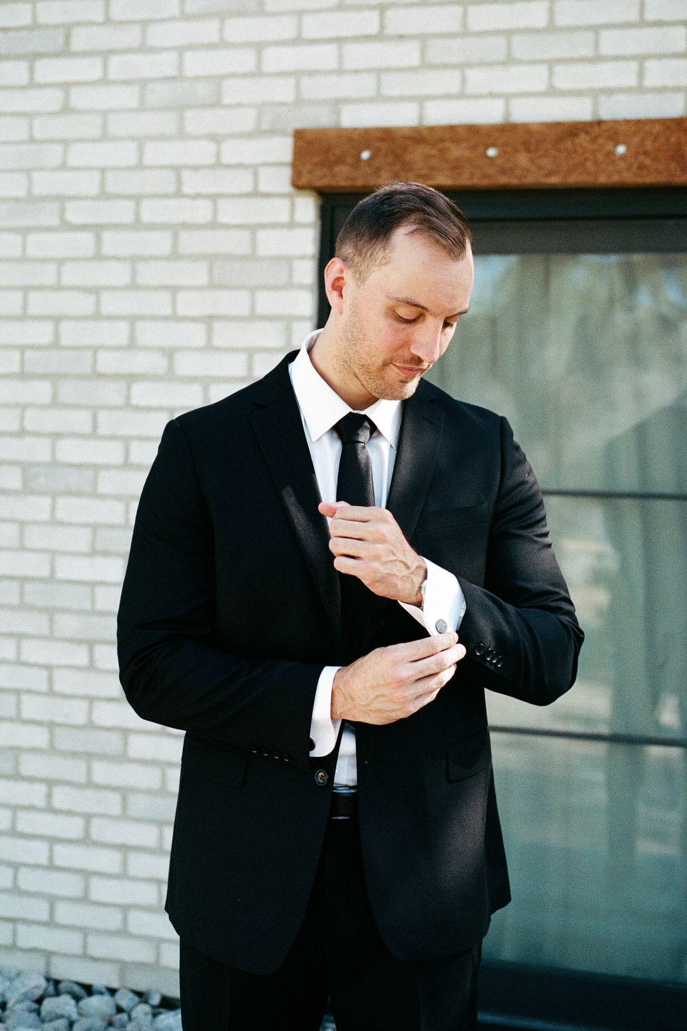 Groom in black tuxedo adjusting cufflinks outside against white brick wall.