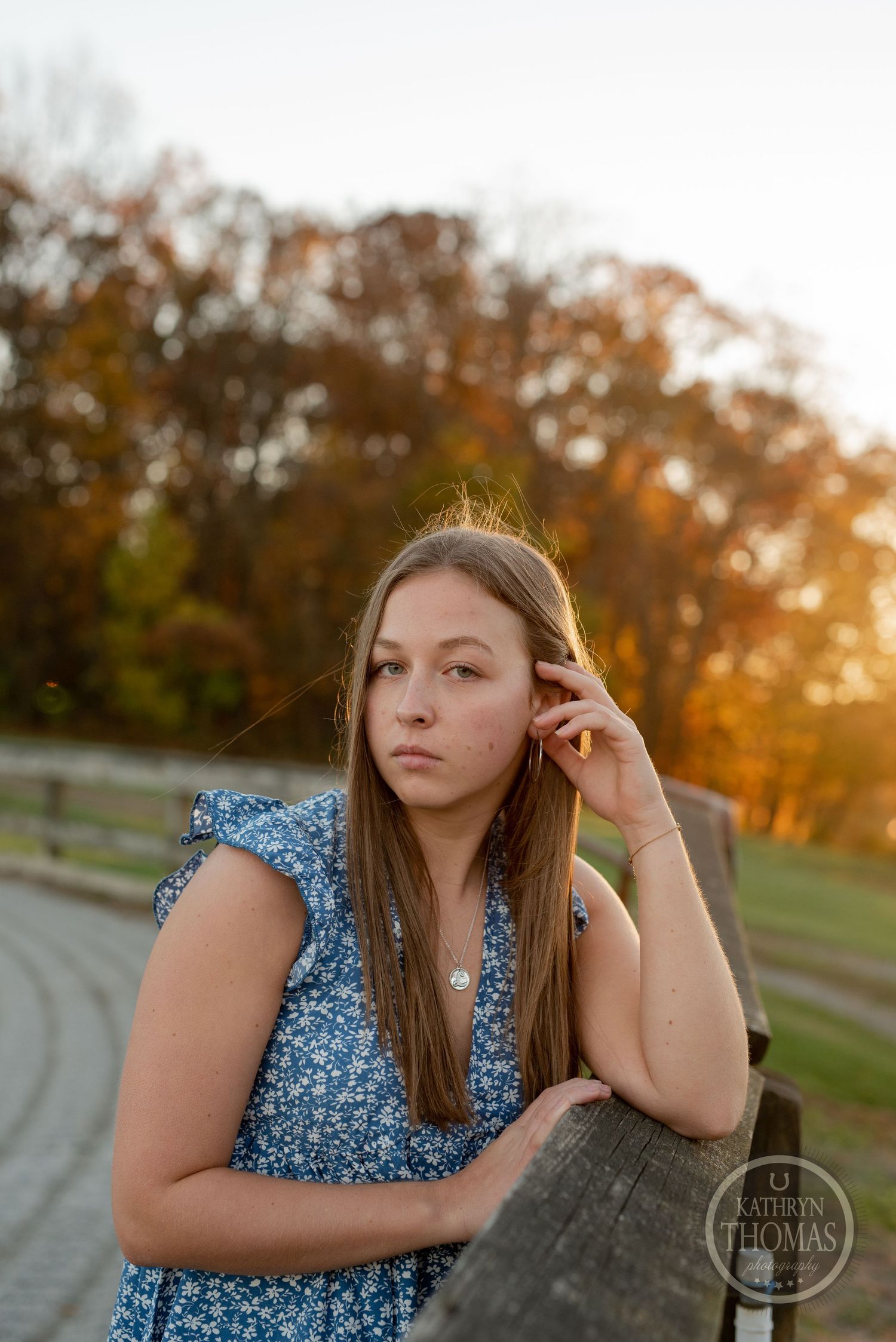 Someone in a blue dress poses thoughtfully by a fence during autumn sunset.