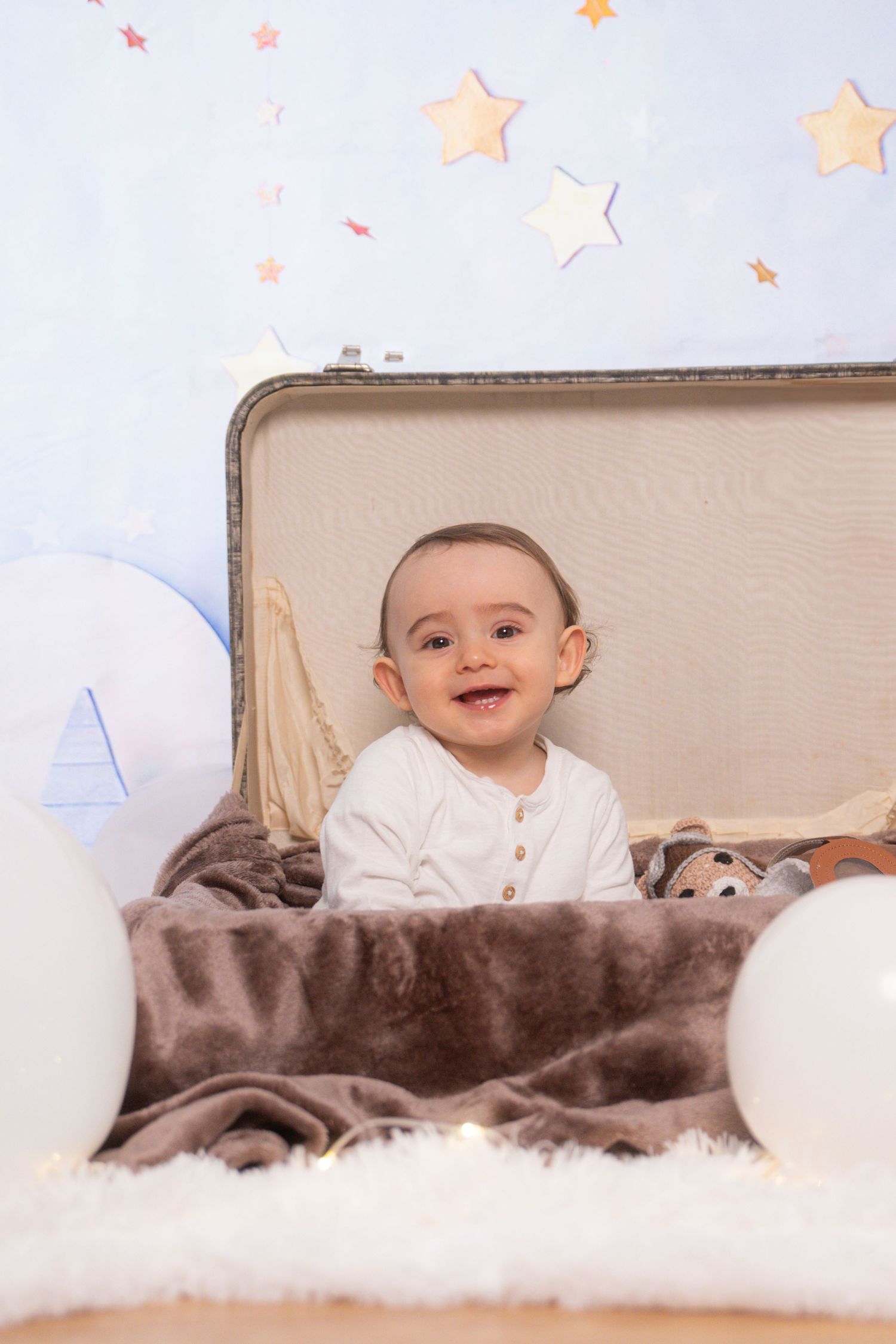 Happy baby peeking from box with plush toys in dreamy star-filled setting.
