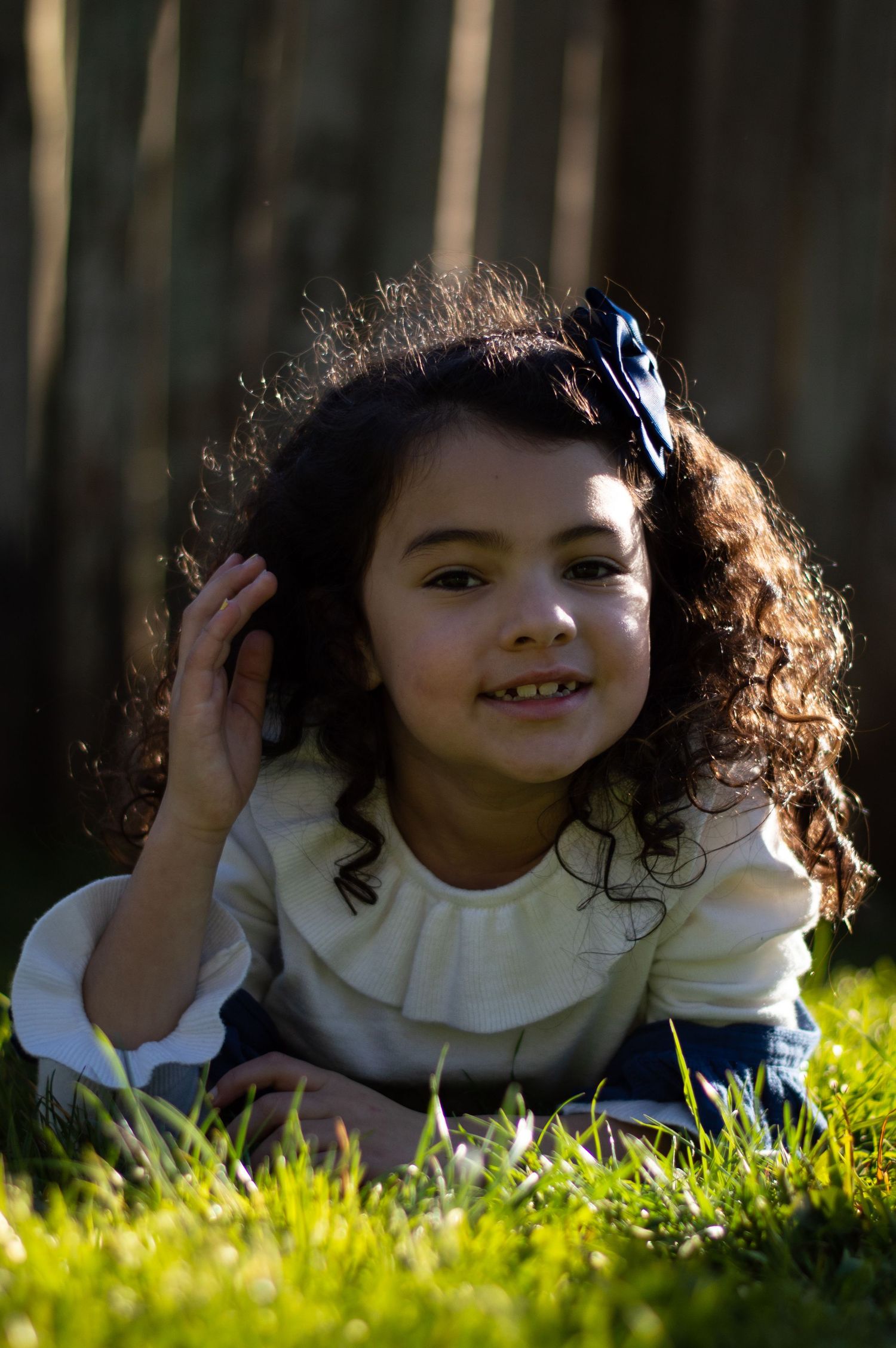 Child with curly hair lying on grass wearing white sweater and hair bow.