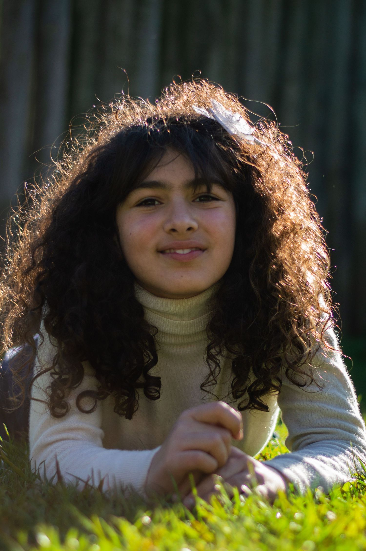 Person with curly hair in cream turtleneck sweater lying on grass in sunlight.