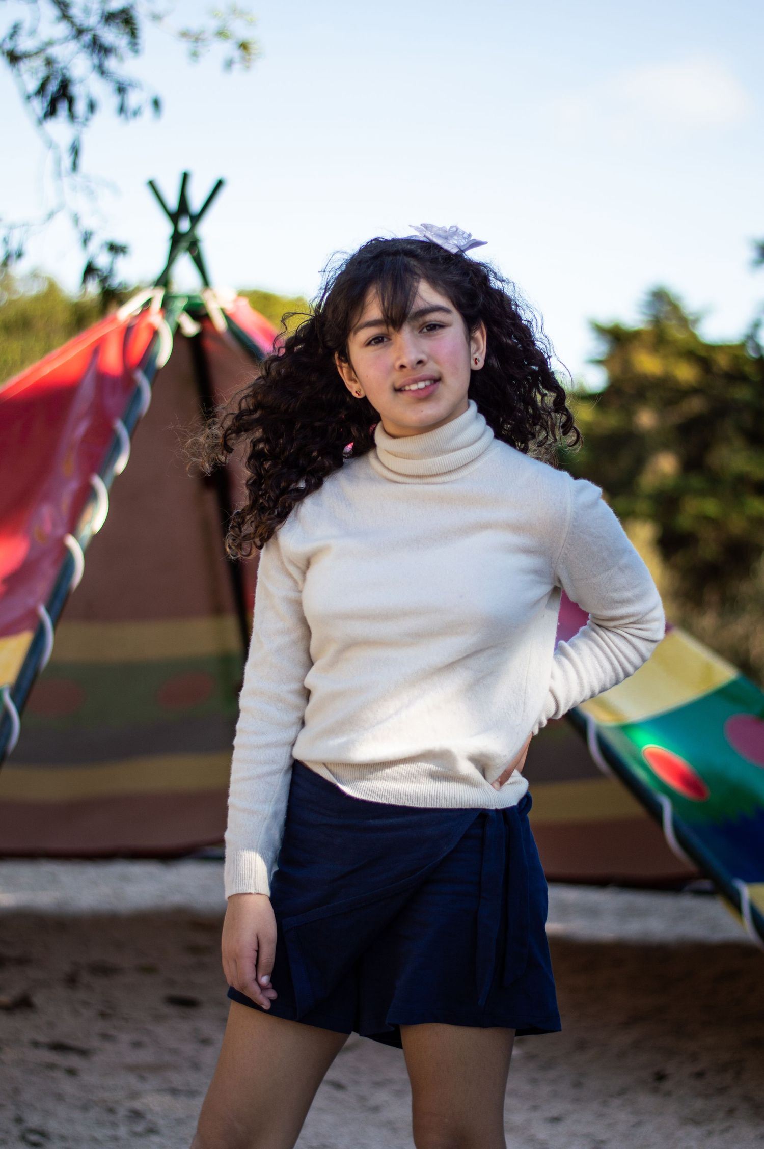 A person in a white turtleneck and navy skirt poses outdoors near playground equipment.