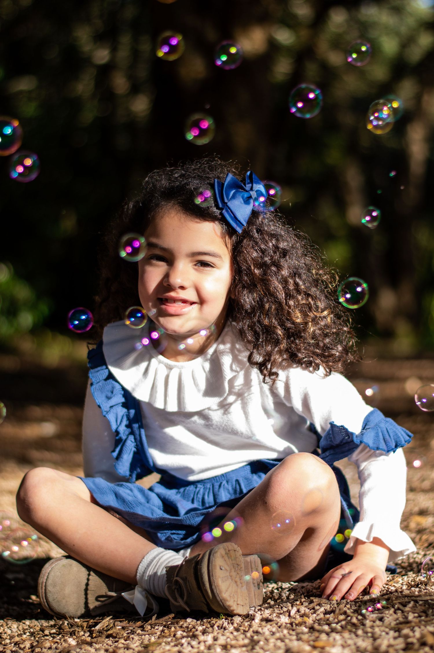 A child in a blue and white dress sits cross-legged outdoors while colorful soap bubbles float in the air.