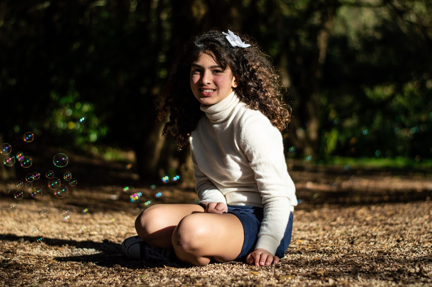 Person in white turtleneck sitting on ground surrounded by sunlit trees.
