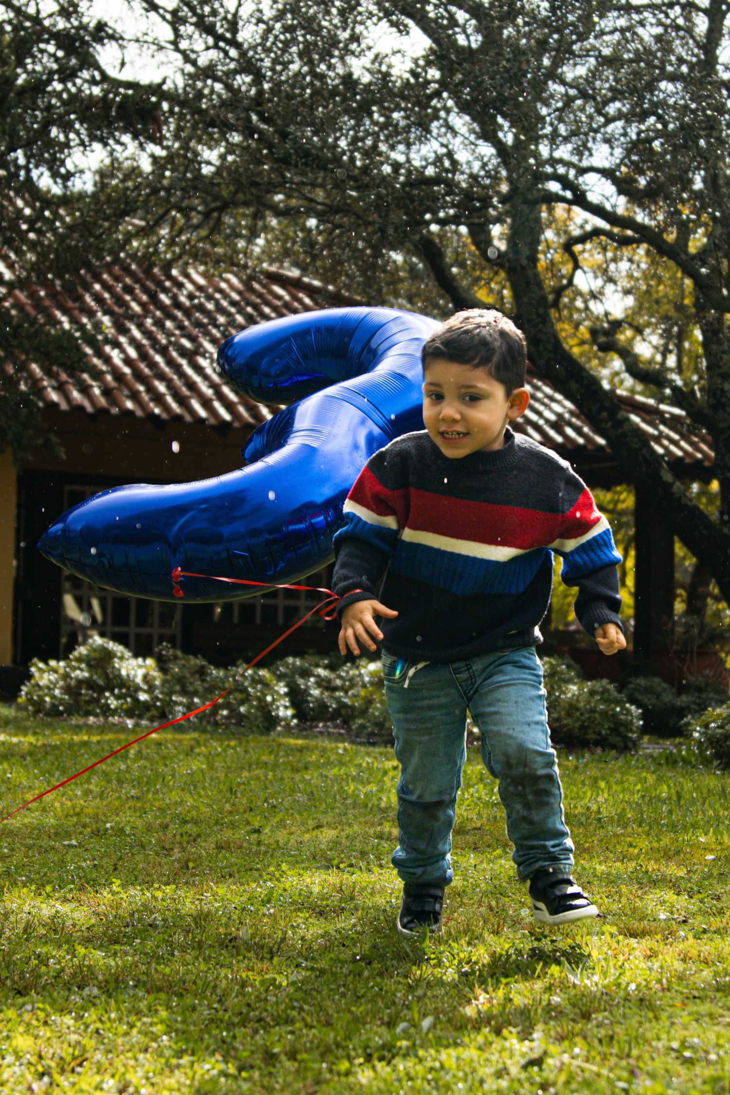 Child in blue jacket and jeans plays energetically in a sunny garden setting.