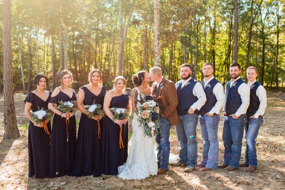 Wedding party poses in forest clearing with bridesmaids in navy dresses and groomsmen in rustic attire.