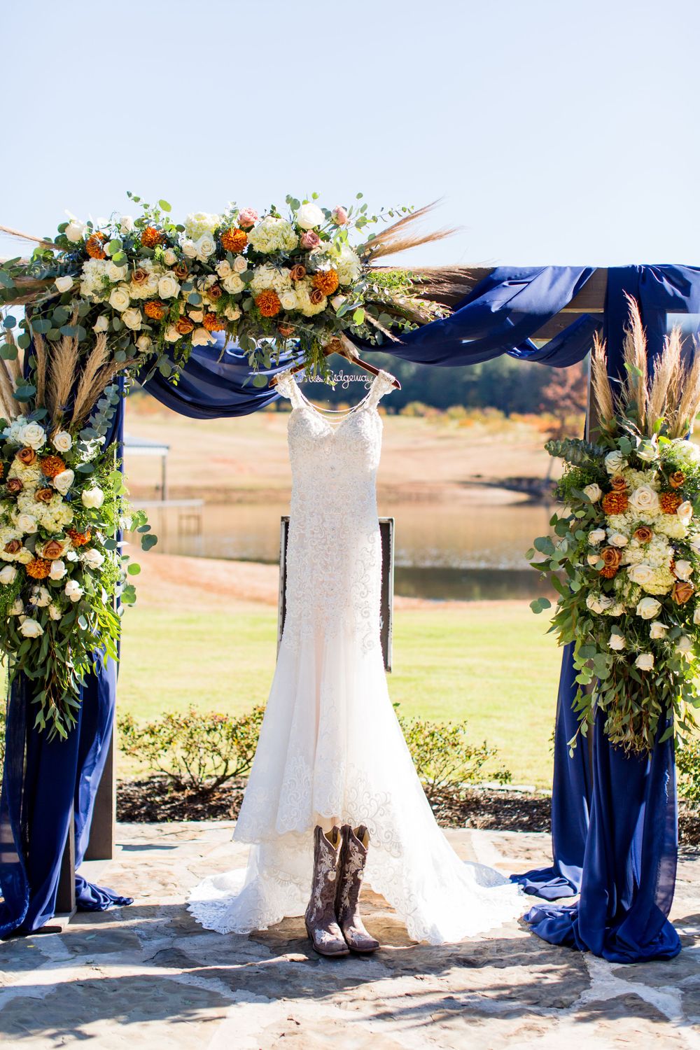 White wedding dress hangs on floral arch with navy blue draping and cowboy boots beneath.