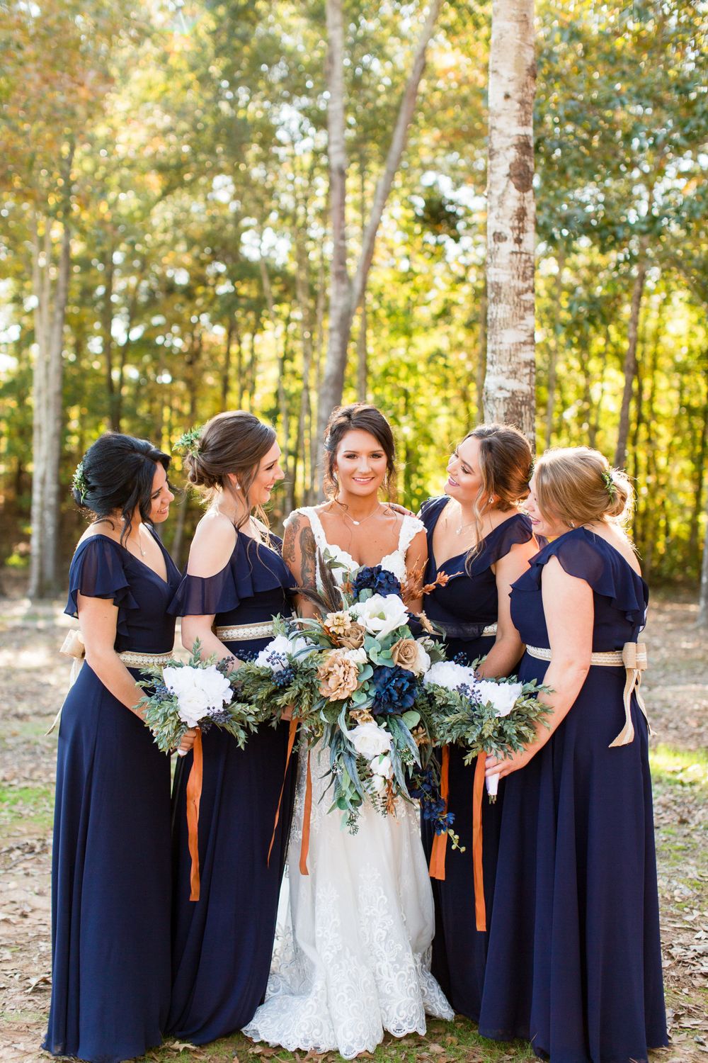 Bridesmaids in navy blue dresses gather around bride holding blue and white floral bouquets in wooded setting.