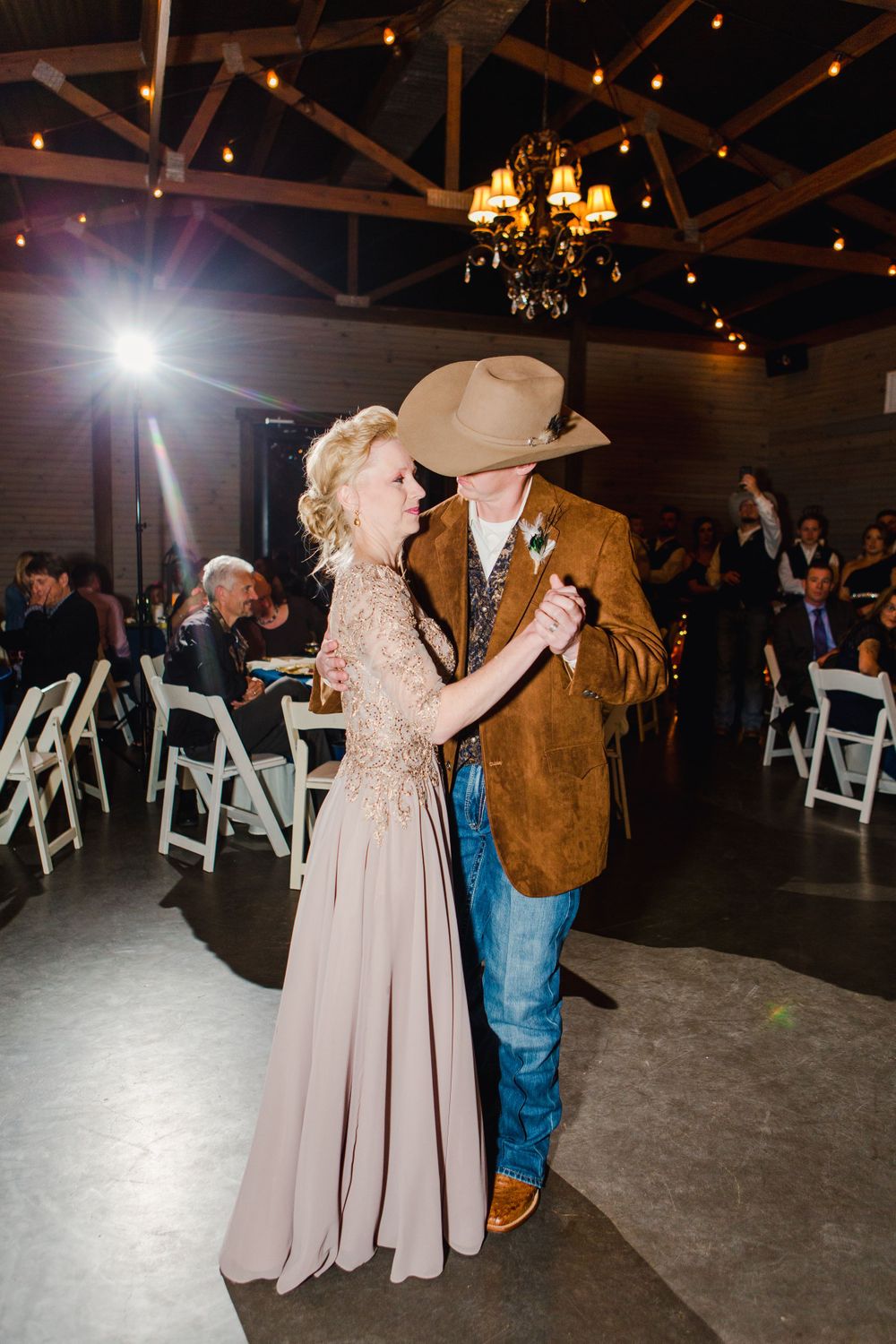 A couple dances intimately at a wedding reception under a rustic chandelier.