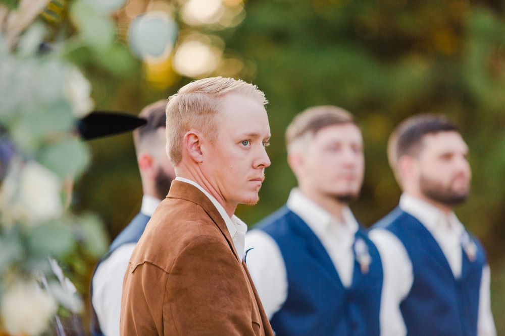 Groomsmen in blue vests stand together during an outdoor autumn wedding ceremony.