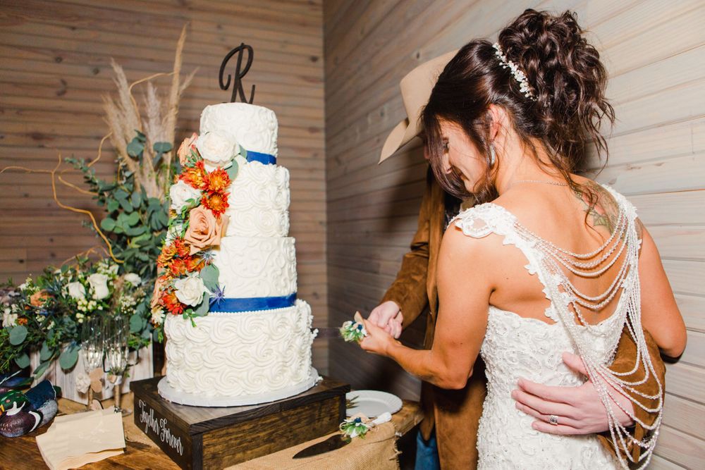 A white wedding cake with blue accents and orange floral decorations stands beside lush greenery.
