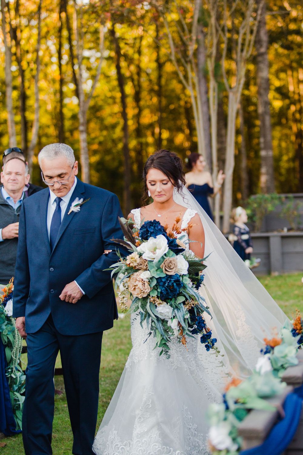 Wedding ceremony in a fall forest setting with beautiful bridal bouquet featuring navy and white flowers.