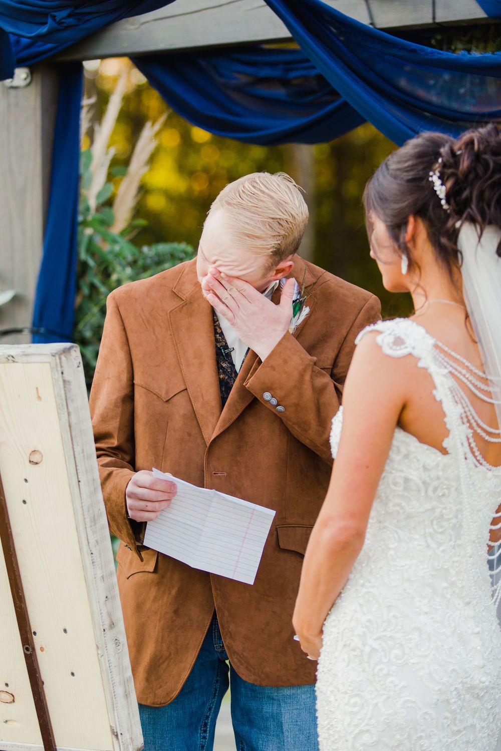 Emotional wedding ceremony moment under blue draped arch with fall foliage in background.