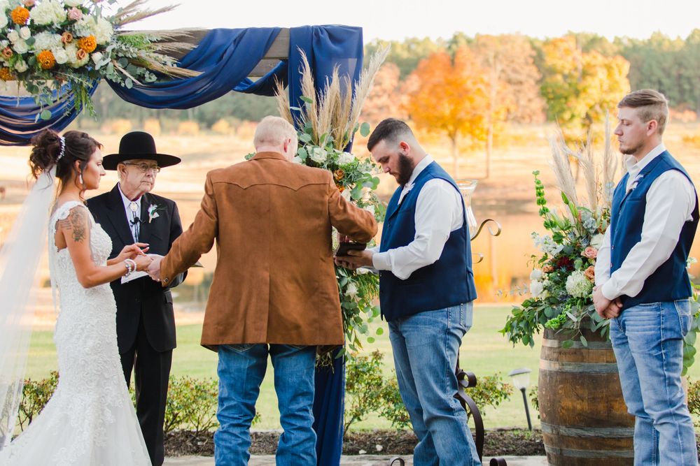 Wedding ceremony under floral arch with navy draping and rustic barrel decor.