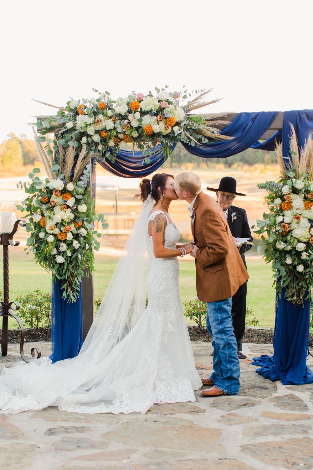 First kiss moment under floral arch with navy draping and orange blooms at outdoor wedding ceremony.