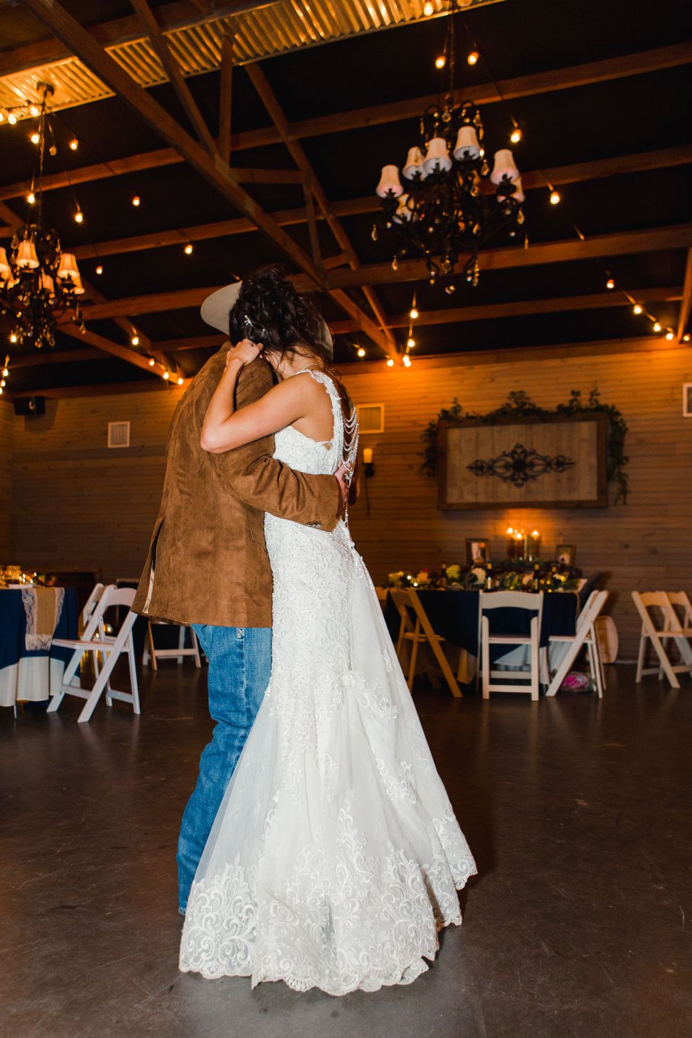 A couple sharing an intimate dance in a rustic reception hall with chandeliers and white folding chairs.