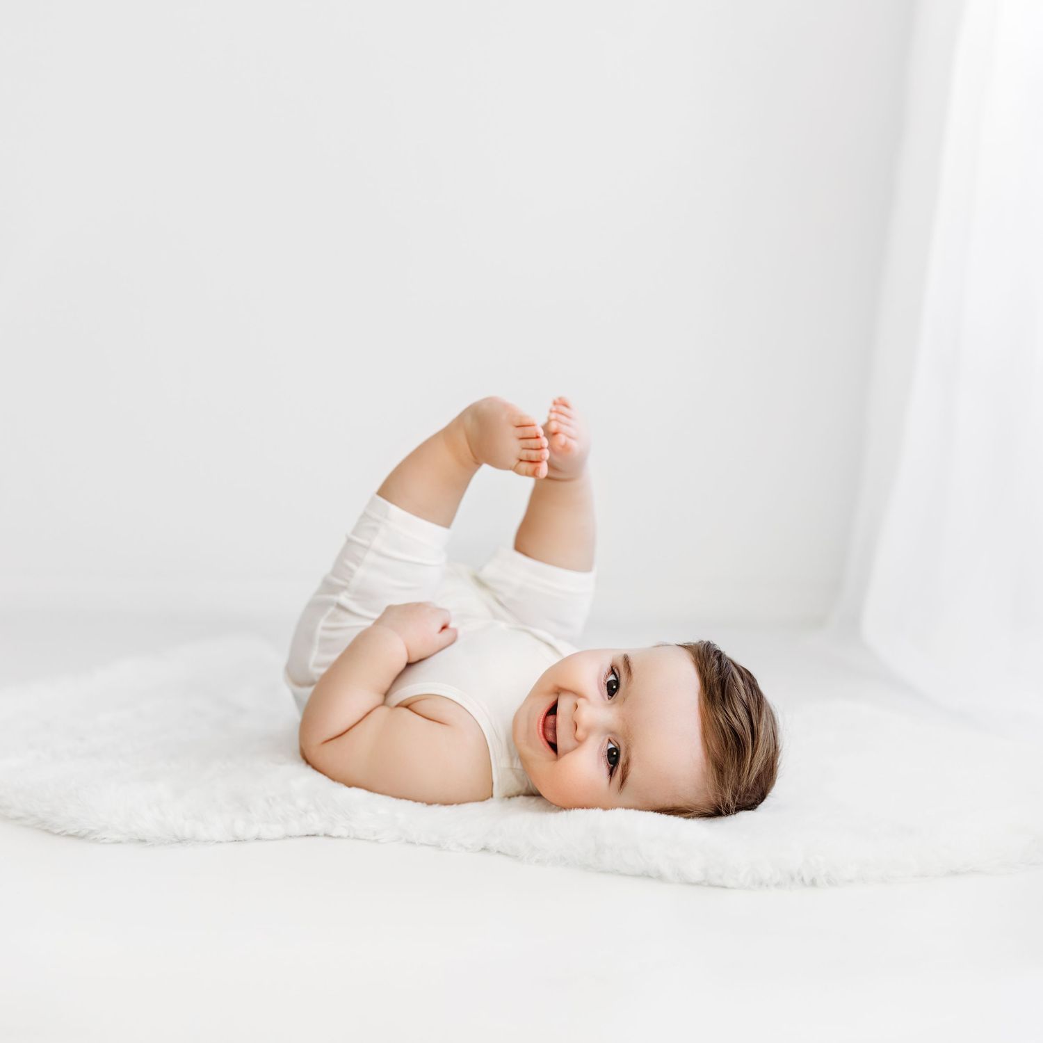 Baby boy laying on white fur rug, feet in air, smiling at camera