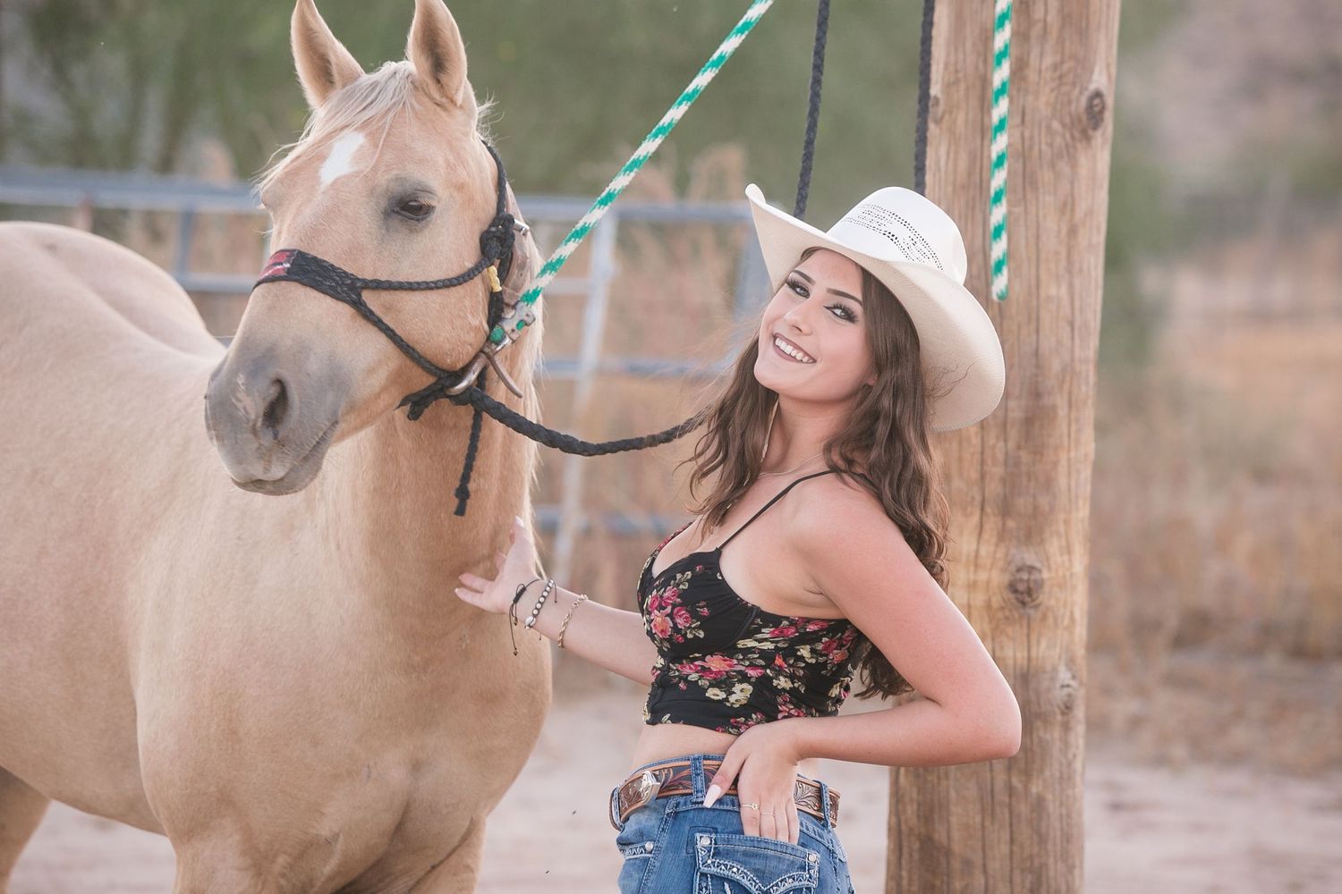 Cheerful woman in white cowboy hat and floral top standing beside a palomino horse outdoors.