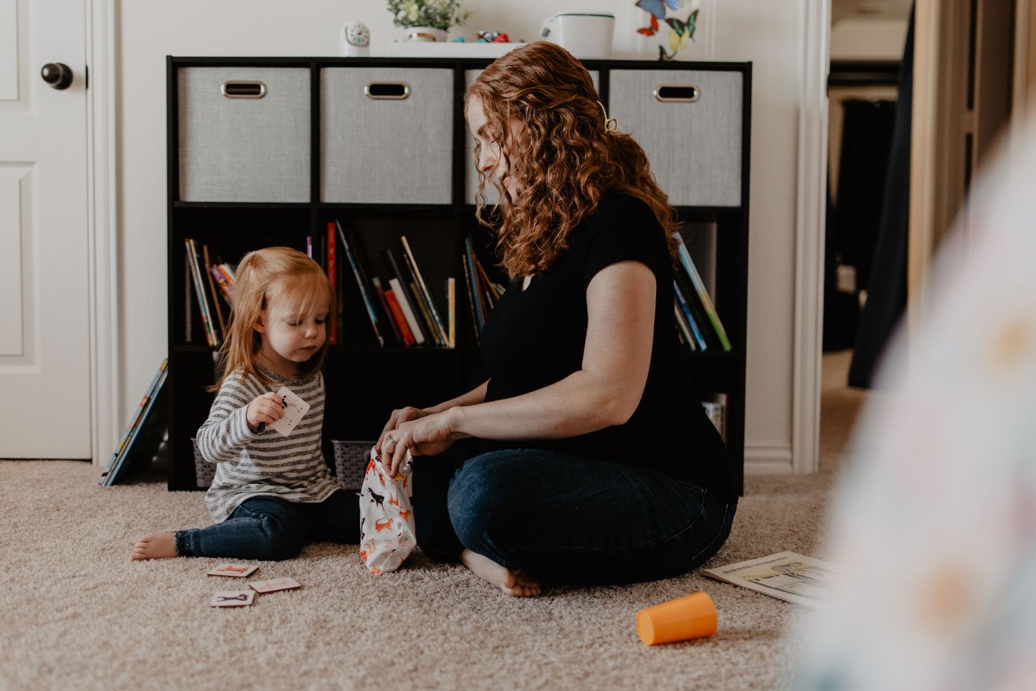 A parent and child play together on carpet near bookshelves and storage bins.