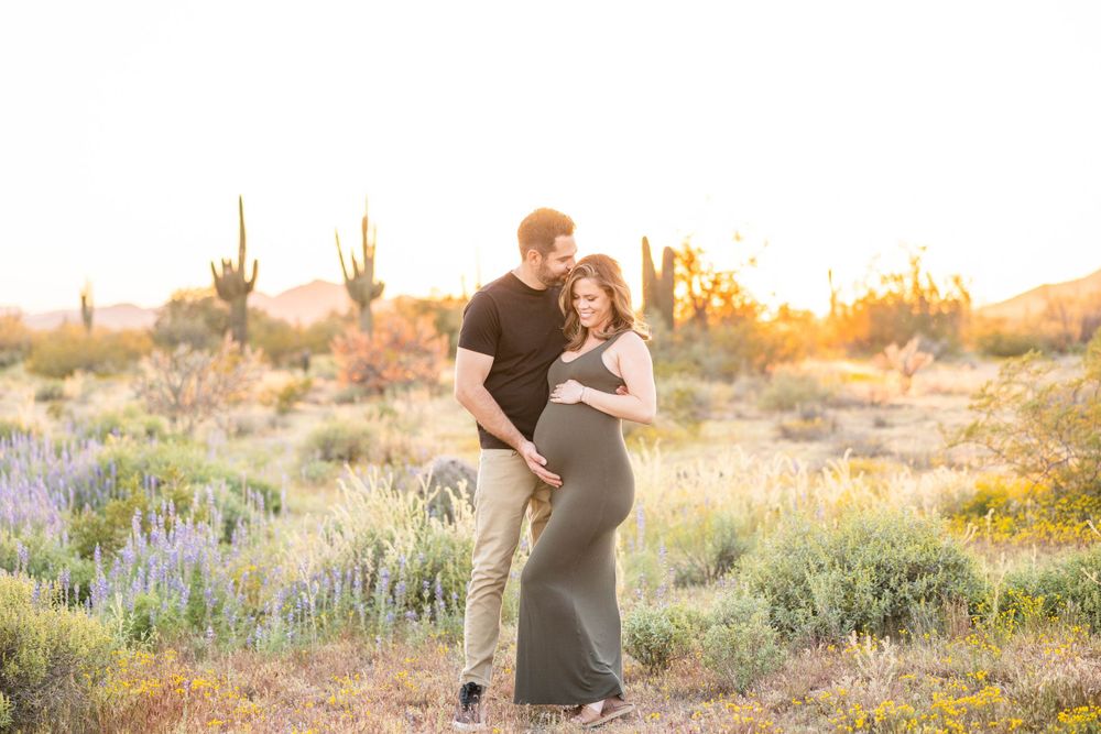 Expecting couple embracing in desert setting with saguaro cacti at sunset.