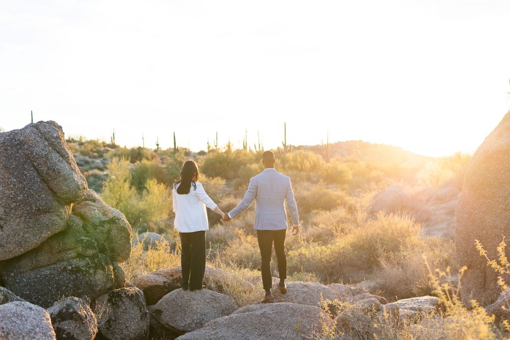 Couple holding hands while walking on rocky terrain during a desert sunset.