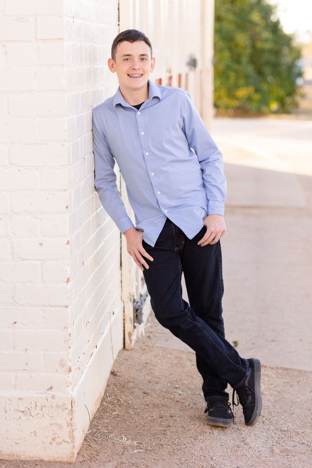 Casual portrait of young man in blue button-down shirt leaning against white wall outdoors.