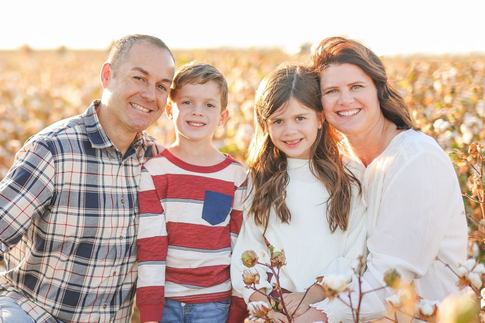 Family of four smiles warmly at camera while posed in cotton field during golden hour.