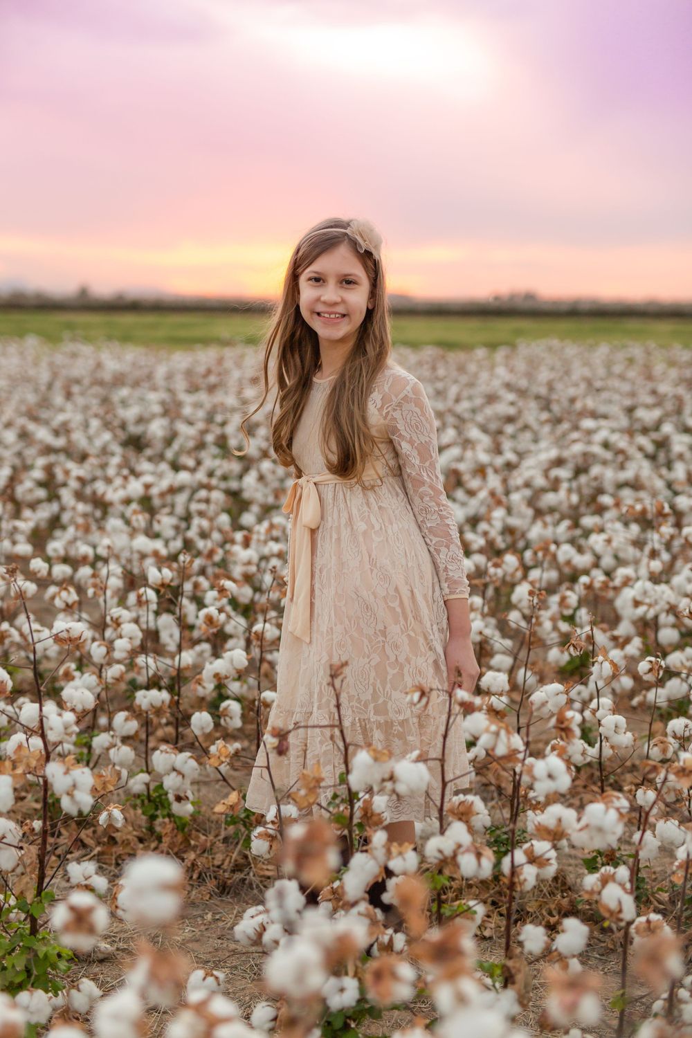 A person in a beige dress stands among fluffy cotton plants during a pink sunset.