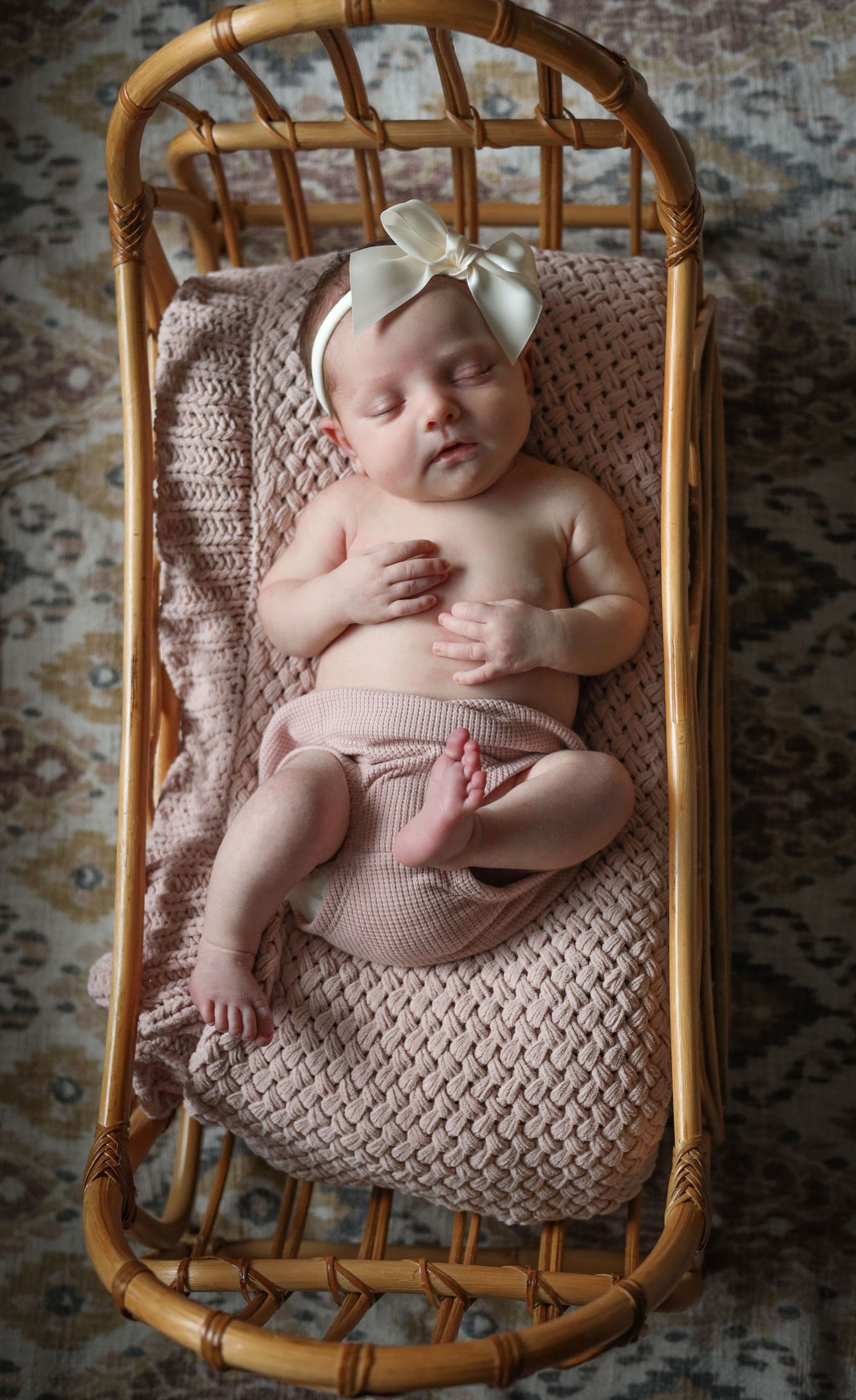 Sleeping newborn in vintage rattan chair with soft knit blanket and delicate bow.