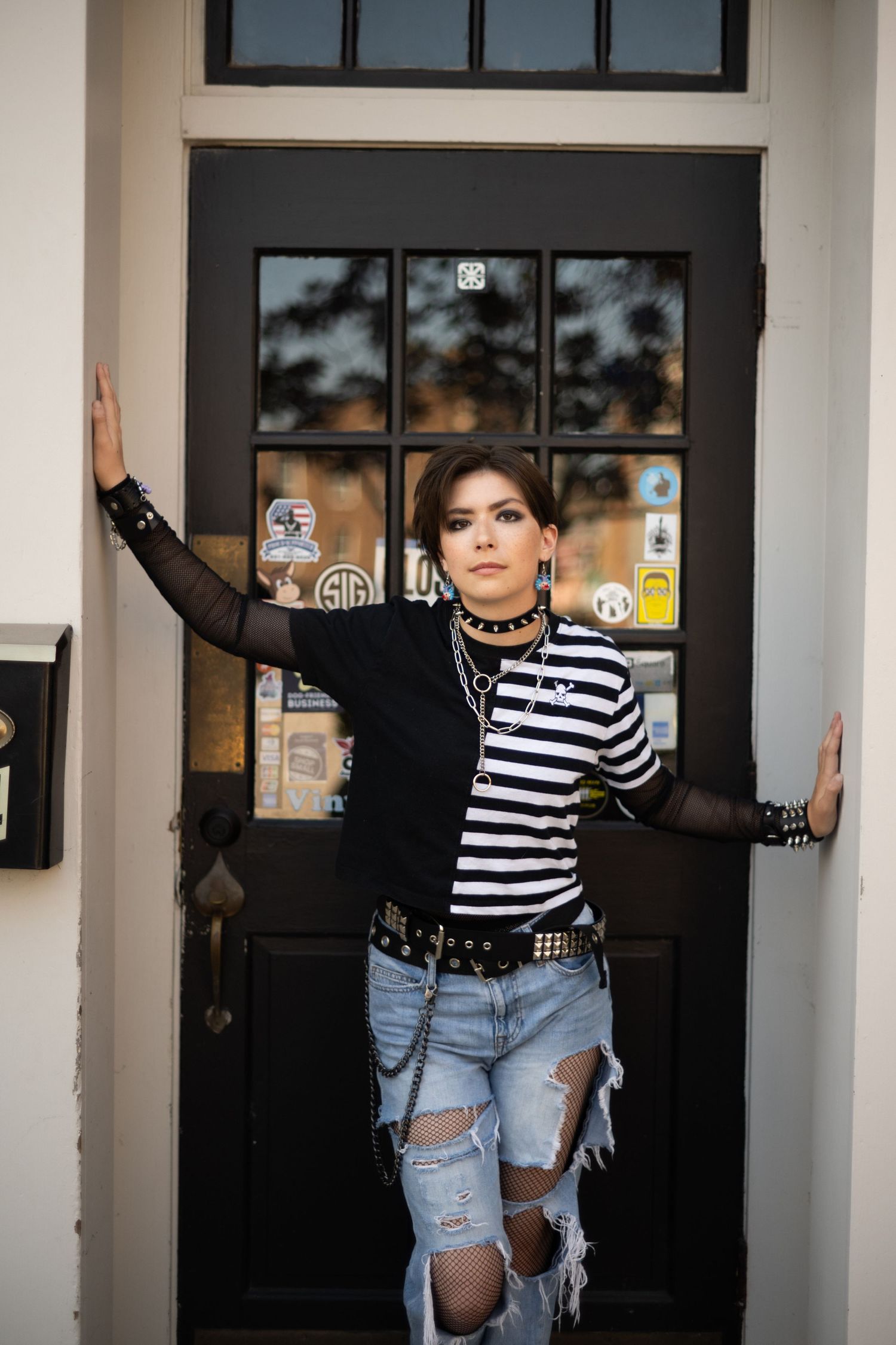 Alternative fashion look with striped shirt, ripped jeans, and punk accessories against a glass door.